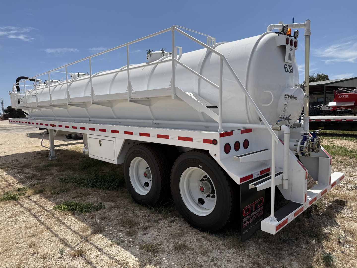 Tank trailer shown from rear-left side with white cylindrical tank on dual-axle chassis, railing along top, rear access steps, and red/white safety stripes.
