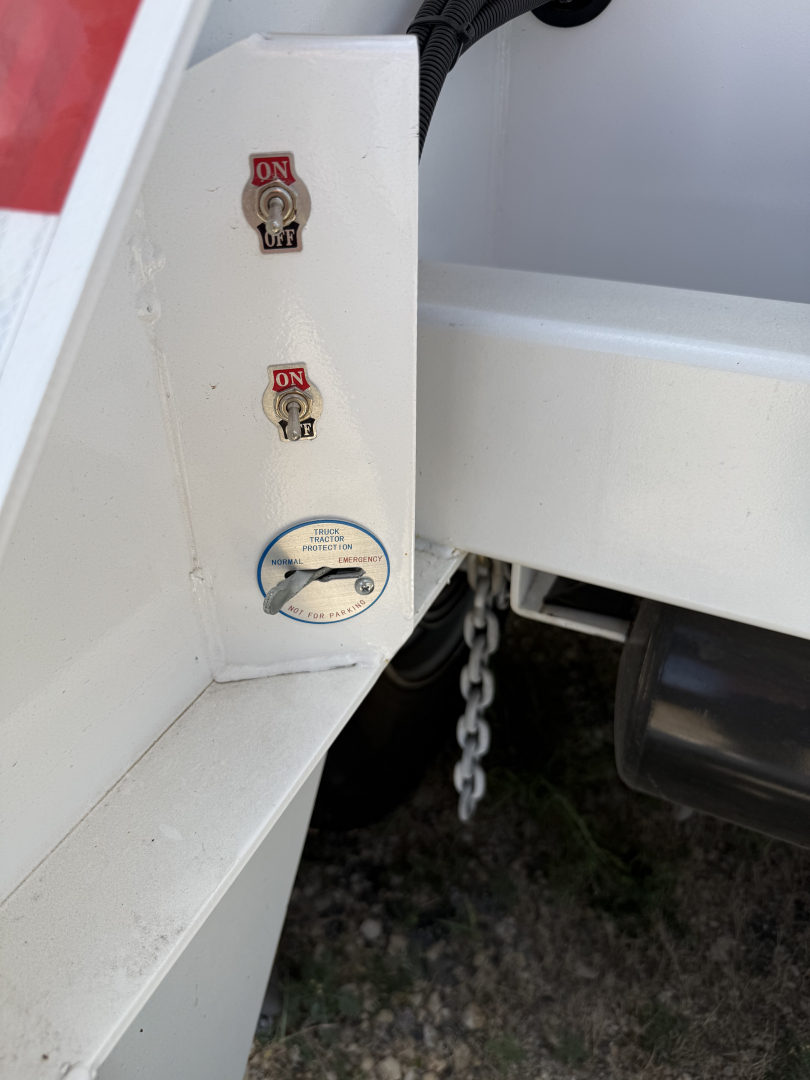 Trailer hitch area showing two red-on-white ON switches and a circular blue-labeled inspection cap near a chain, with white metal frame.