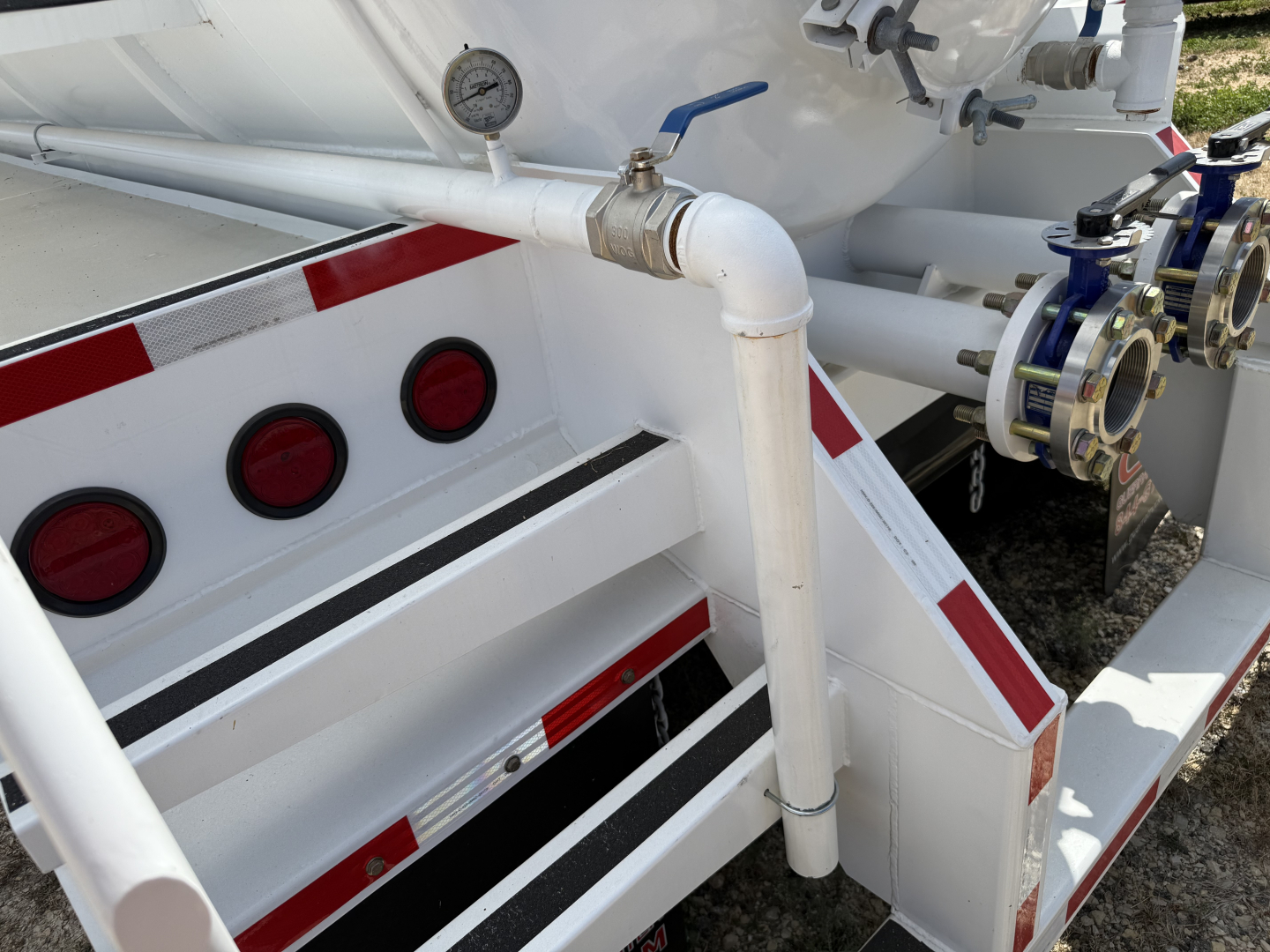 Vacuum trailer rear showing piping, gauge, and valve assembly on white tank with red reflective stripes