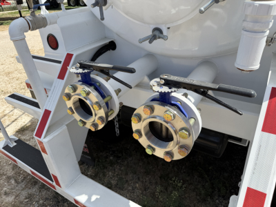 Two large metal flanges with bolted flanges and valves on a tanker trailer rear, hoses connected, with black handles and blue gaskets; white painted metal frame and red reflective stripes.