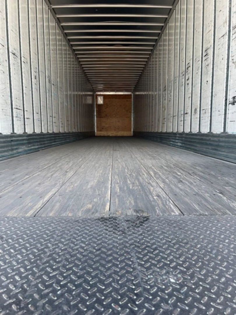 Rear interior of a utility van trailer with metal walls and wooden floor, empty cargo area, diamond plate at rear threshold.