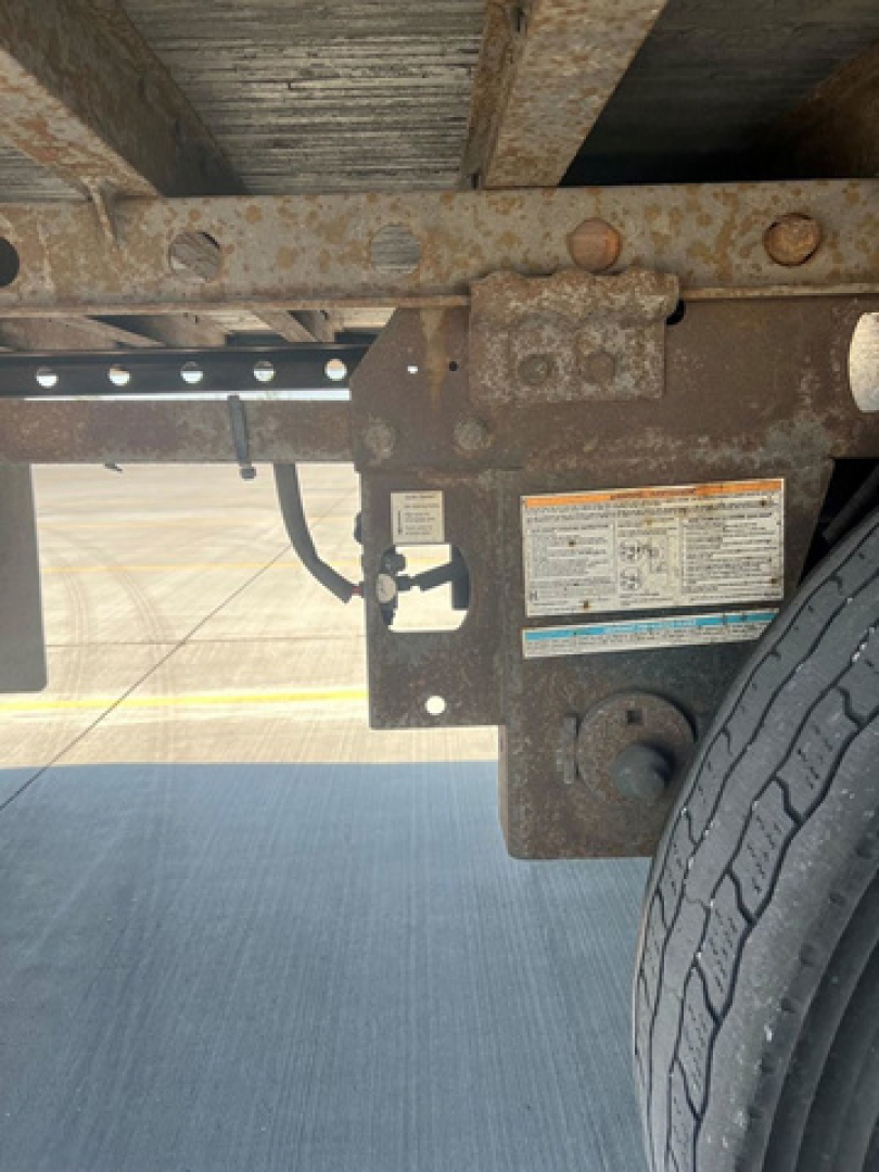 Trailer chassis undercarriage showing wheel, frame rails, and suspension mounting with rust and a label plate visible.