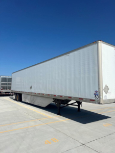 Semi-trailer in a parking lot with no visible cab; box trailer shows plain silver walls with rear undercarriage and landing gear visible.
