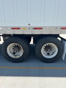 Dual rear wheels of a utility trailer with a white corrugated side, showing riveted panels and wheel rims.
