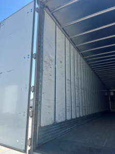 Trailer interior showing metal wall panels and side door of a utility van trailer, empty cargo area with ribbed floor and rear entry.