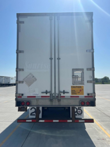 Back of a white utility van trailer showing rear doors with locking bars, taillights, and a CAUTION sticker. Visible text includes 'Glims?' on left door, a small diamond warning placard, and a yellow caution label near bottom right.