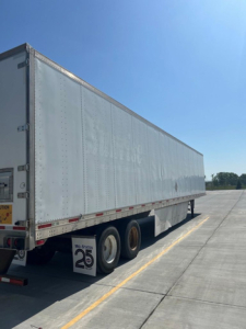 Utility van trailer in a parking lot showing side view of a long white trailer with wheels and a clearance sign.