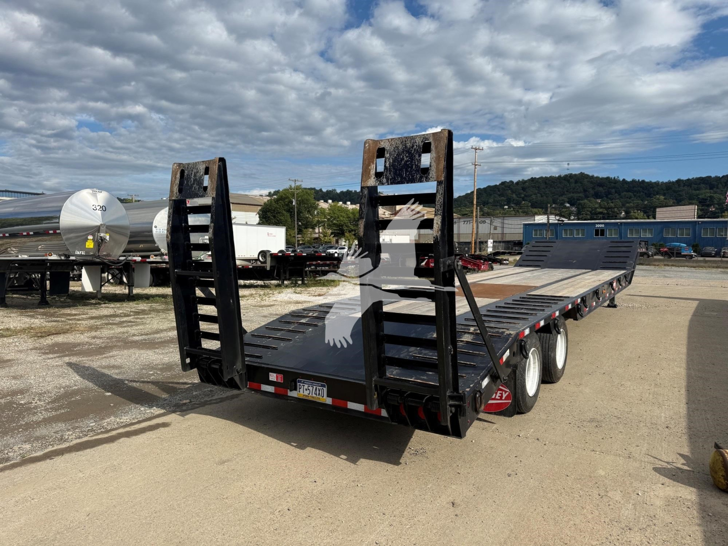 Trailer deck with dual ramps and gooseneck-style rear; black steel with perforated ramps, triple-axle configuration, daylight sky reflection.