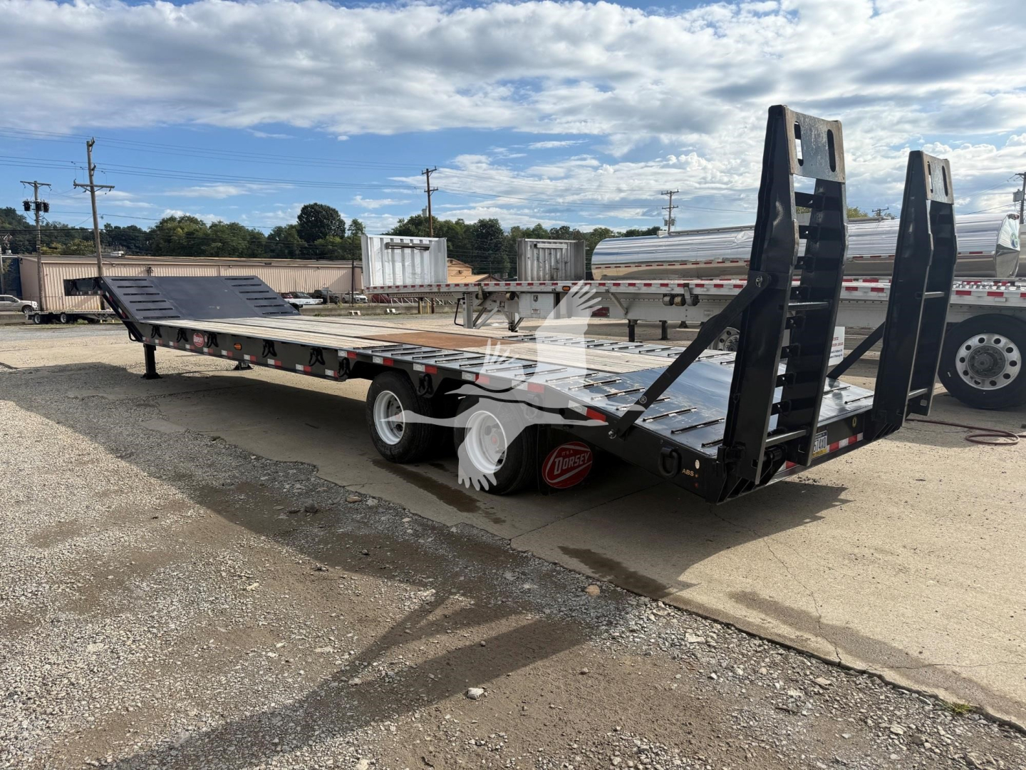 Lowboy trailer with dual ramps and long flat deck showing rear ramps raised, black finish, white wheels, and red wheel chocks; side rails and support beams visible.