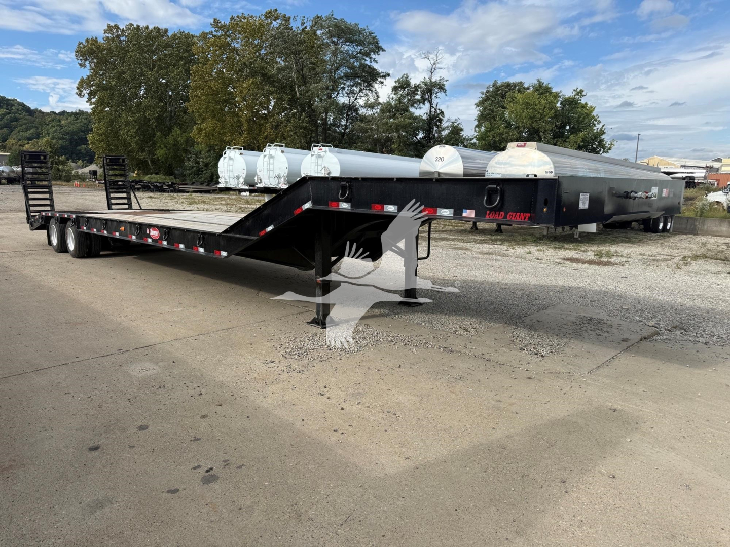 Lowboy trailer with long flatbed and gooseneck, showing dual axles and loading ramps; some white cylindrical tanks in background; no legible brand markings visible on the trailer.