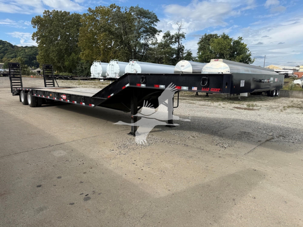Lowboy trailer with long flatbed and gooseneck, showing dual axles and loading ramps; some white cylindrical tanks in background; no legible brand markings visible on the trailer.