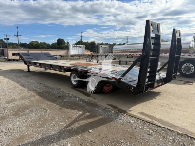 Lowboy trailer with dual ramps and long flat deck showing rear ramps raised, black finish, white wheels, and red wheel chocks; side rails and support beams visible.