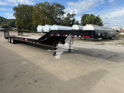Lowboy trailer with long flatbed and gooseneck, showing dual axles and loading ramps; some white cylindrical tanks in background; no legible brand markings visible on the trailer.
