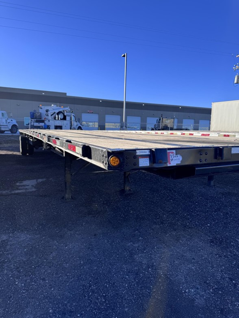 Flatbed trailer showing steel deck with tandem axle and slider. Side markers and rear lights visible; flatbed with open space and light gray frame.