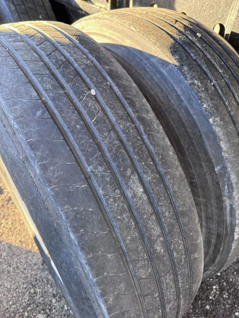 Flatbed trailer wheel close-up showing dual tires with tread and grime; part of steel frame visible in background.