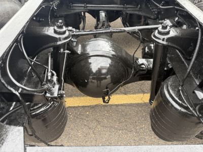 Kenworth T880 rear axle view showing solid differential housing, dual rear tires, suspension components, and drive shaft; black undercarriage with wet surface.
