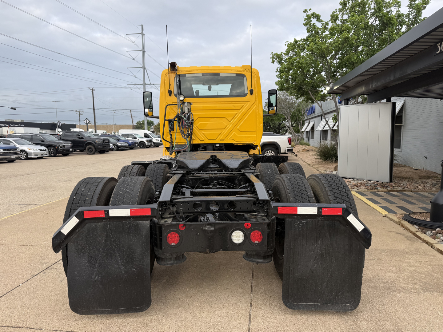 Truck tractor rear view with fifth wheel and dual rear axles, yellow cab, black chassis, mud flaps, and various reflection from dealership lot.