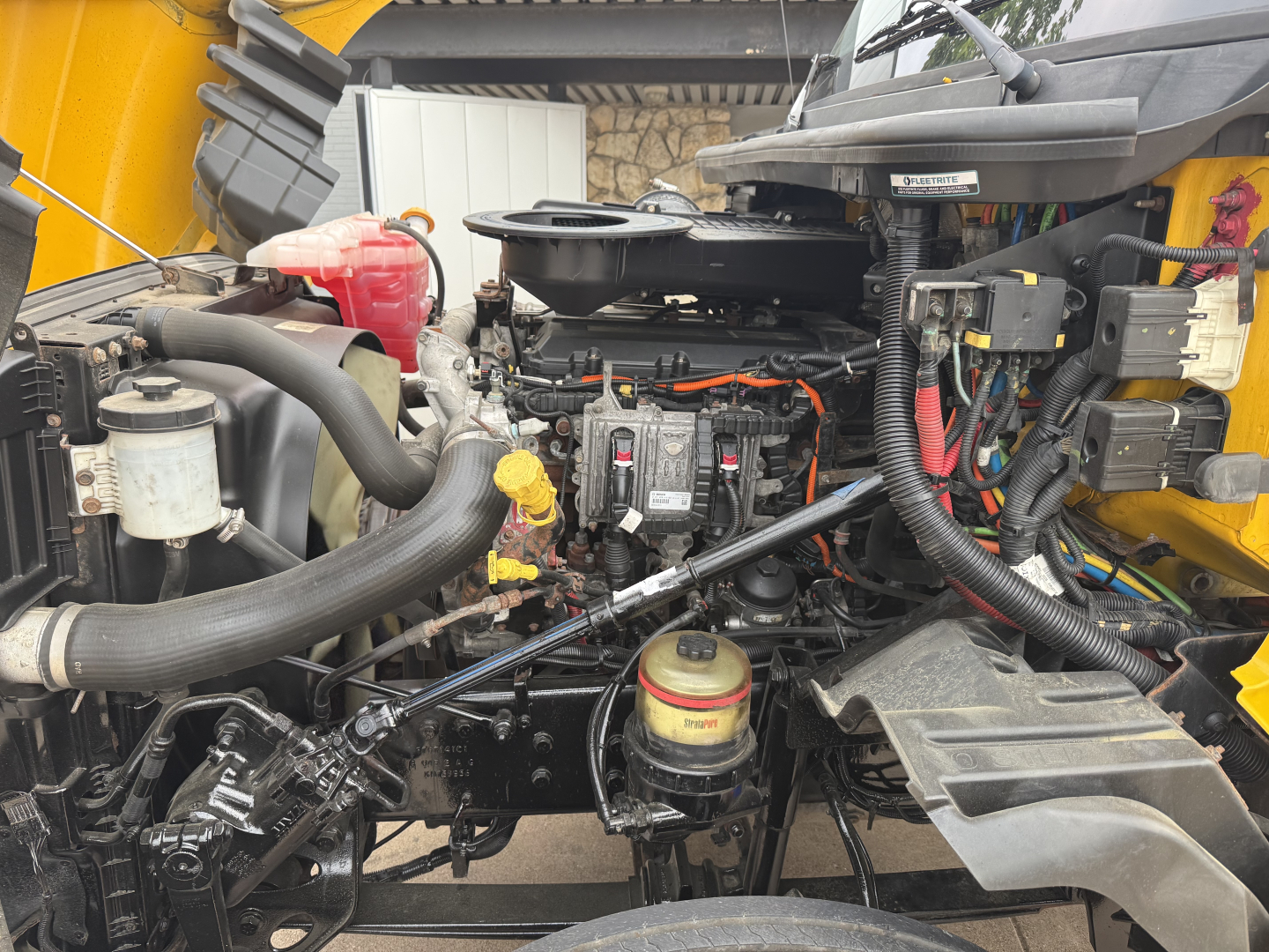 Truck engine bay showing International LT diesel engine with visible coolant reservoir, hoses, wiring harness, and components in a yellow cab.