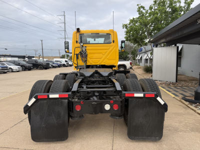 Truck tractor rear view with fifth wheel and dual rear axles, yellow cab, black chassis, mud flaps, and various reflection from dealership lot.