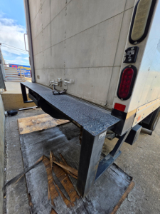 Box truck rear view with folding steel step and taillights visible on right; raised loading platform and wood debris on ground.