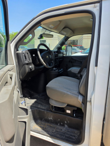 Front interior of a 2016 Chevrolet Express 3500 cargo van showing driver's seat, steering wheel, dashboard, and open entry with black floor mat.