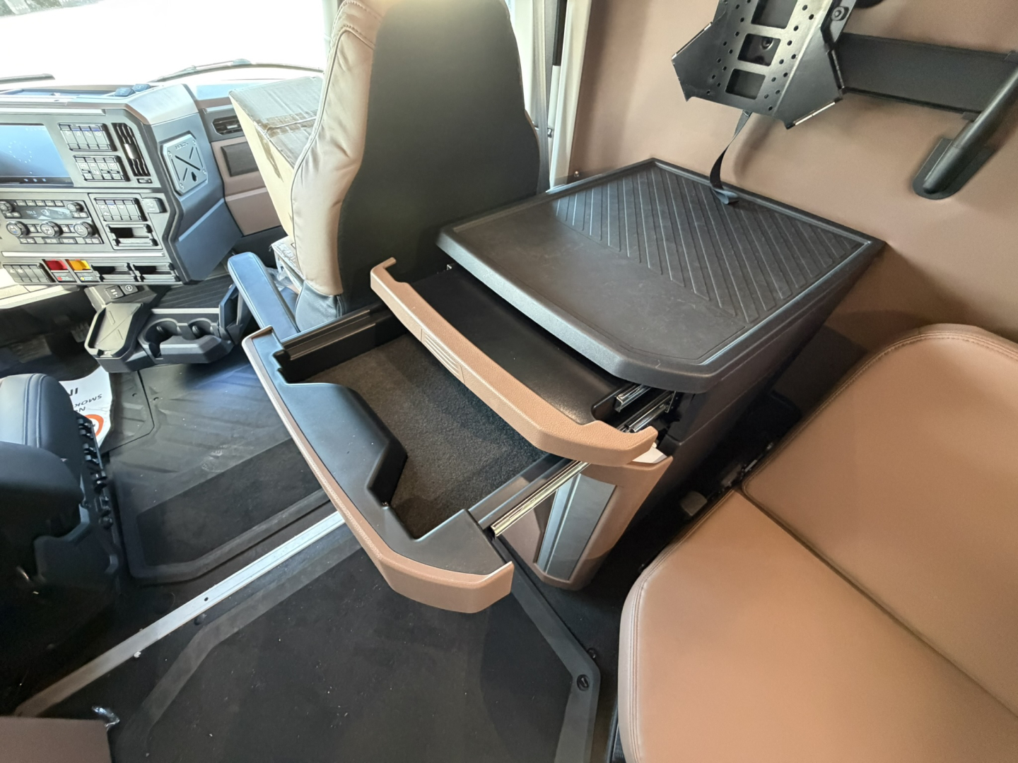 Cab interior of a Mack truck showing an open center console with storage drawers and a tray, beige seats and dashboard visible.