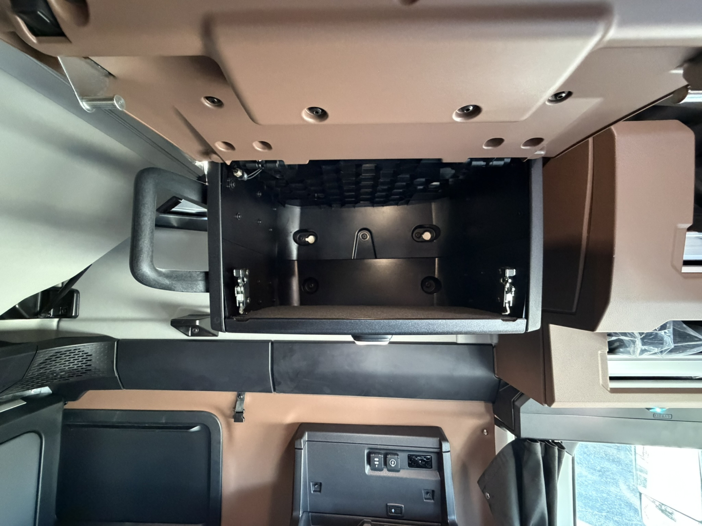 Truck interior storage compartment under dash in a Mack PR64T76H showing an open empty box with mounting brackets and screws.