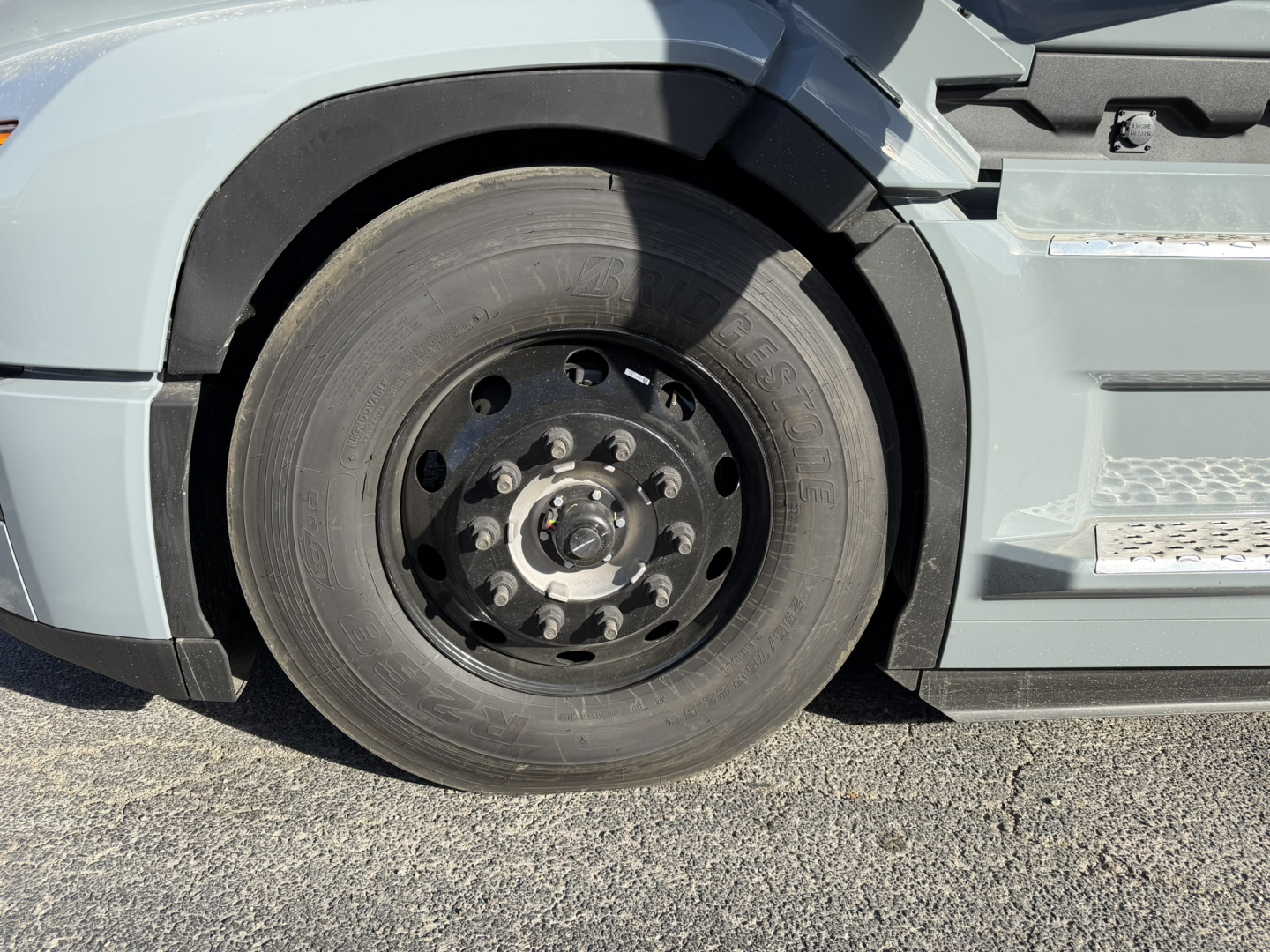 Truck front wheel area showing large black steel wheel with 10 lug nuts and tire, part of Mack PR64T76H cabover, side step and fender visible