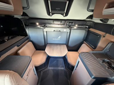 Truck cab interior showing lounge seating and table, Mack logo on rear cabinet, brown leatherette upholstery, overhead controls, and center table.