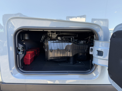 Truck engine bay in a Mack PR64T76H showing engine components and battery bank in the front storage area, with access door open.