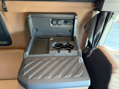 Interior console area of a 2026 Mack PR64T76H showing a center armrest with cup holders and a small storage tray; inset USB/12V power outlets and a control panel on the wall above.