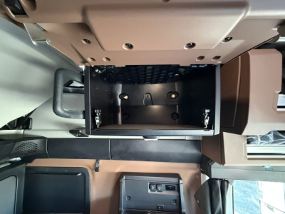 Truck interior storage compartment under dash in a Mack PR64T76H showing an open empty box with mounting brackets and screws.