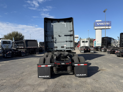 Mack PR64T76H truck tractor back view showing dual rear wheels, black mud flaps with 'MACK' text, trailer connection area, and a dealership lot in the background.