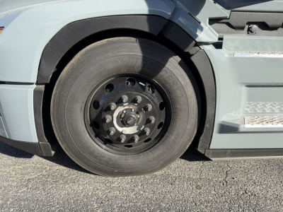 Truck front wheel area showing large black steel wheel with 10 lug nuts and tire, part of Mack PR64T76H cabover, side step and fender visible