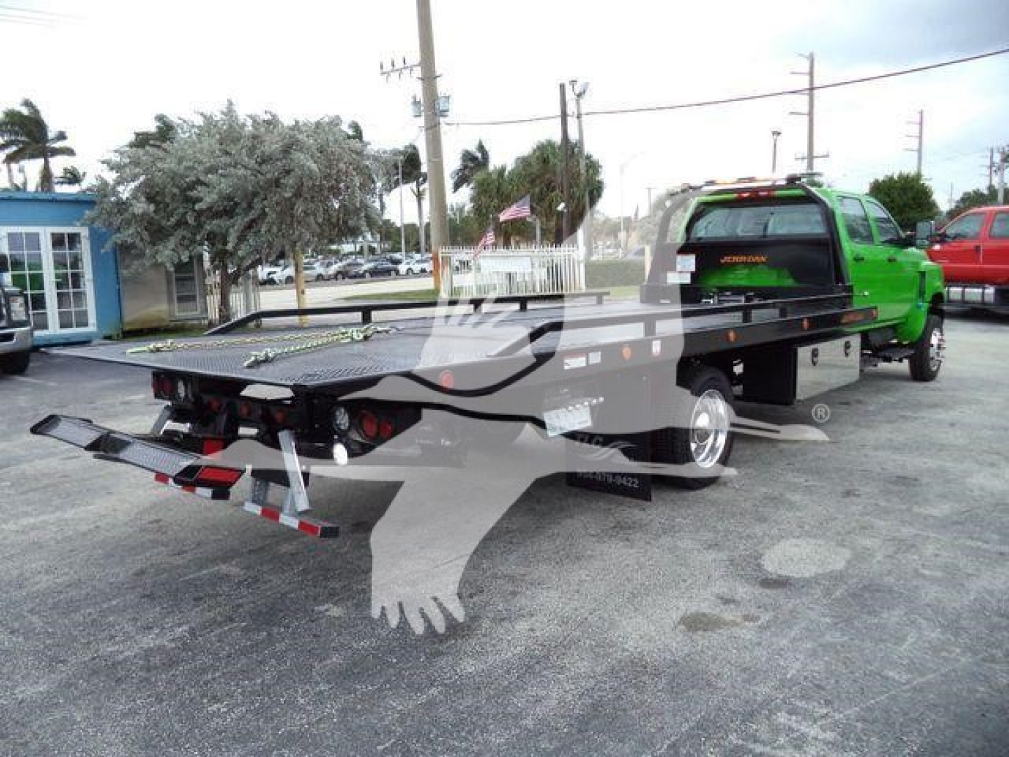 Flatbed rollback tow truck with green cab and black bed, rear view showing taillights, towing hitch, and reflective safety stripes on the bumper.