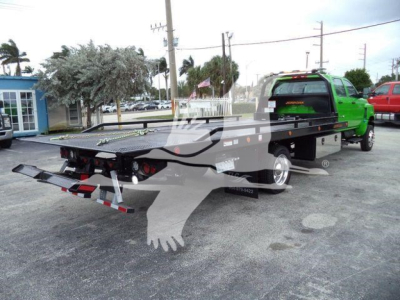 Flatbed rollback tow truck with green cab and black bed, rear view showing taillights, towing hitch, and reflective safety stripes on the bumper.