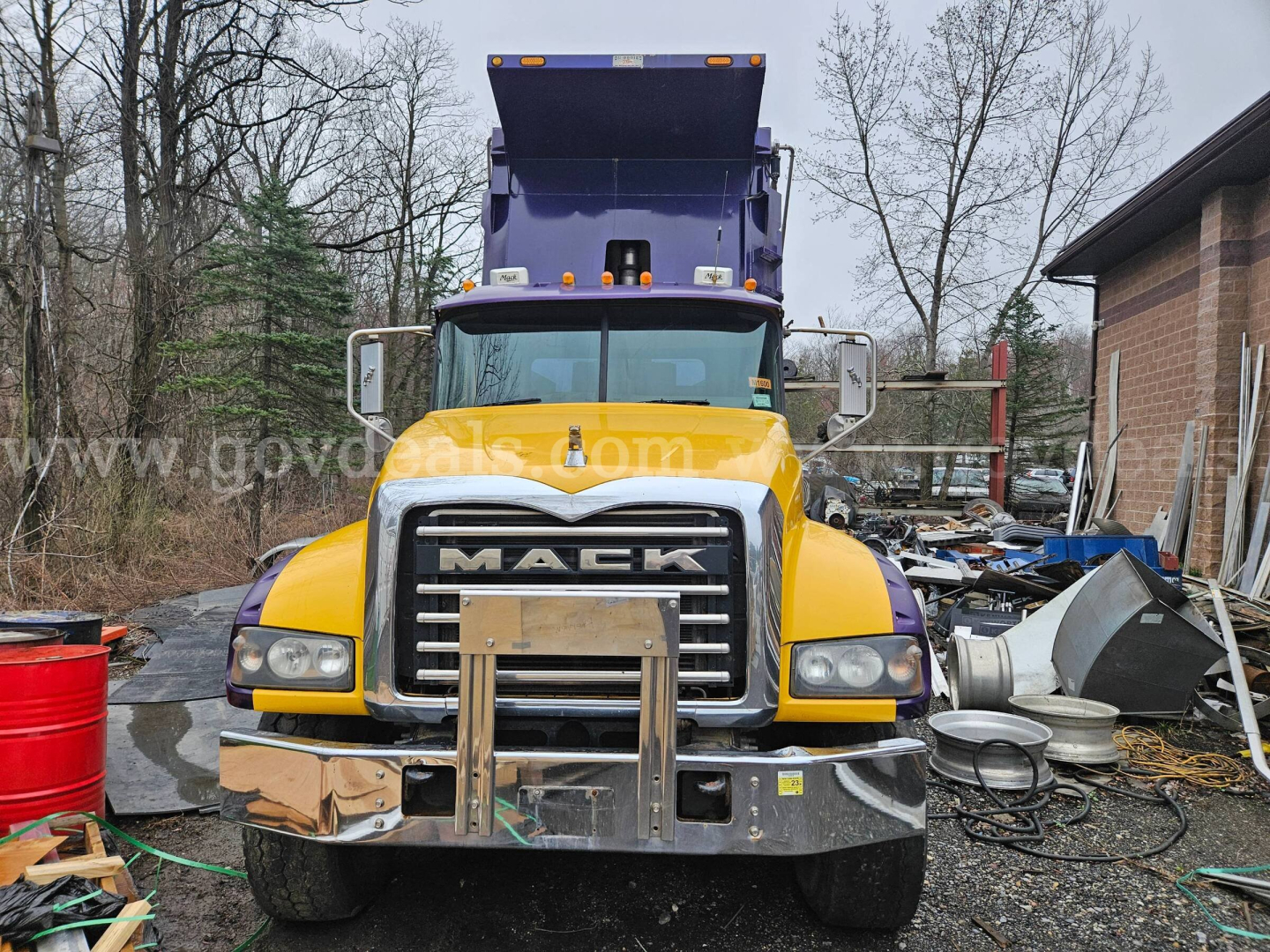 Front view of a 2014 Mack GU713 dump truck with yellow cab, chrome grille, purple dump bed raised overhead, parked in a lot with spare parts around.
