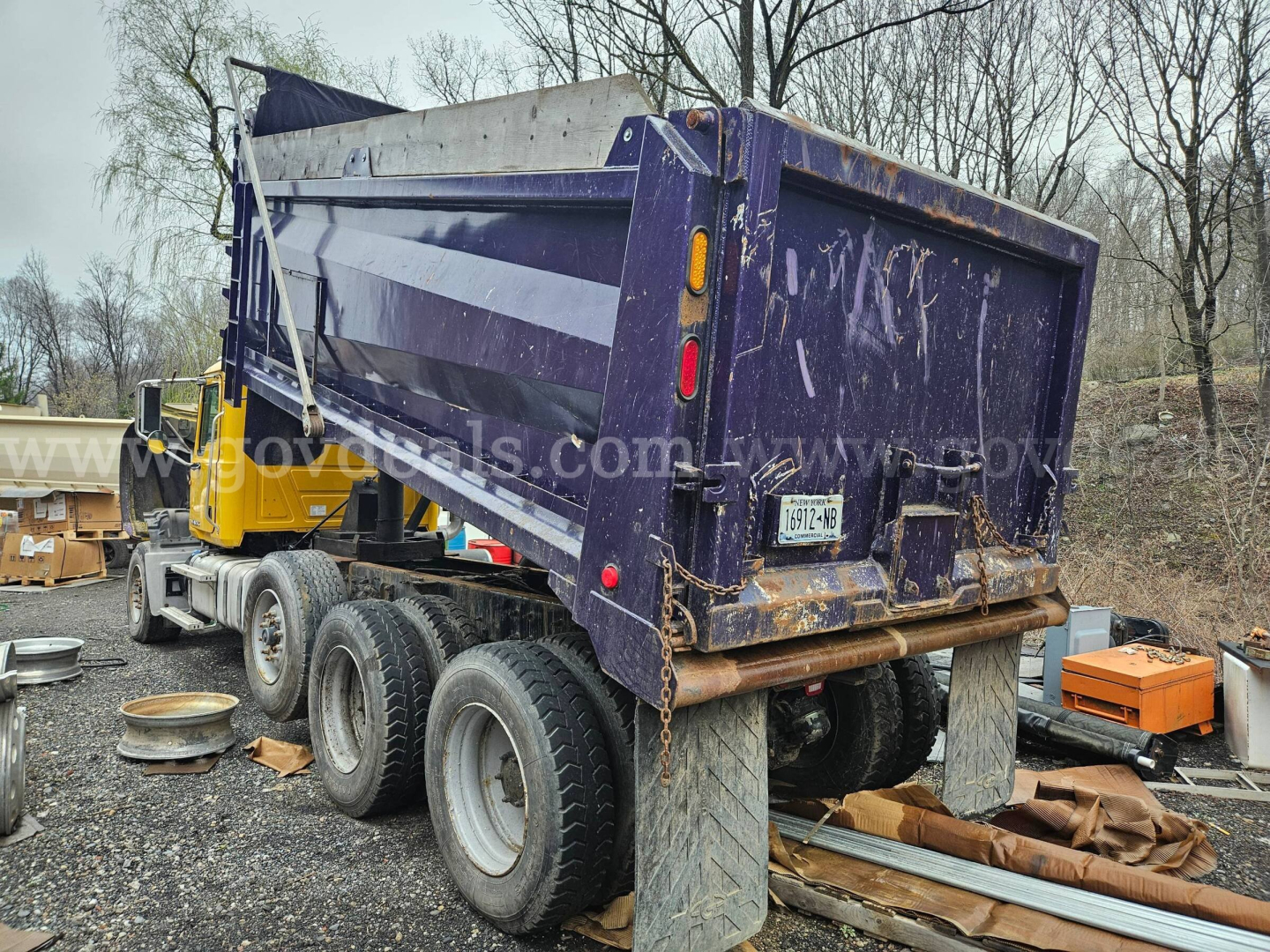 Dump truck with purple bed tilted up, rear view showing tailgate, mud flaps, and triple-axle wheels; yard with equipment and trees in background.