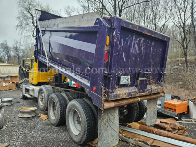 Dump truck with purple bed tilted up, rear view showing tailgate, mud flaps, and triple-axle wheels; yard with equipment and trees in background.