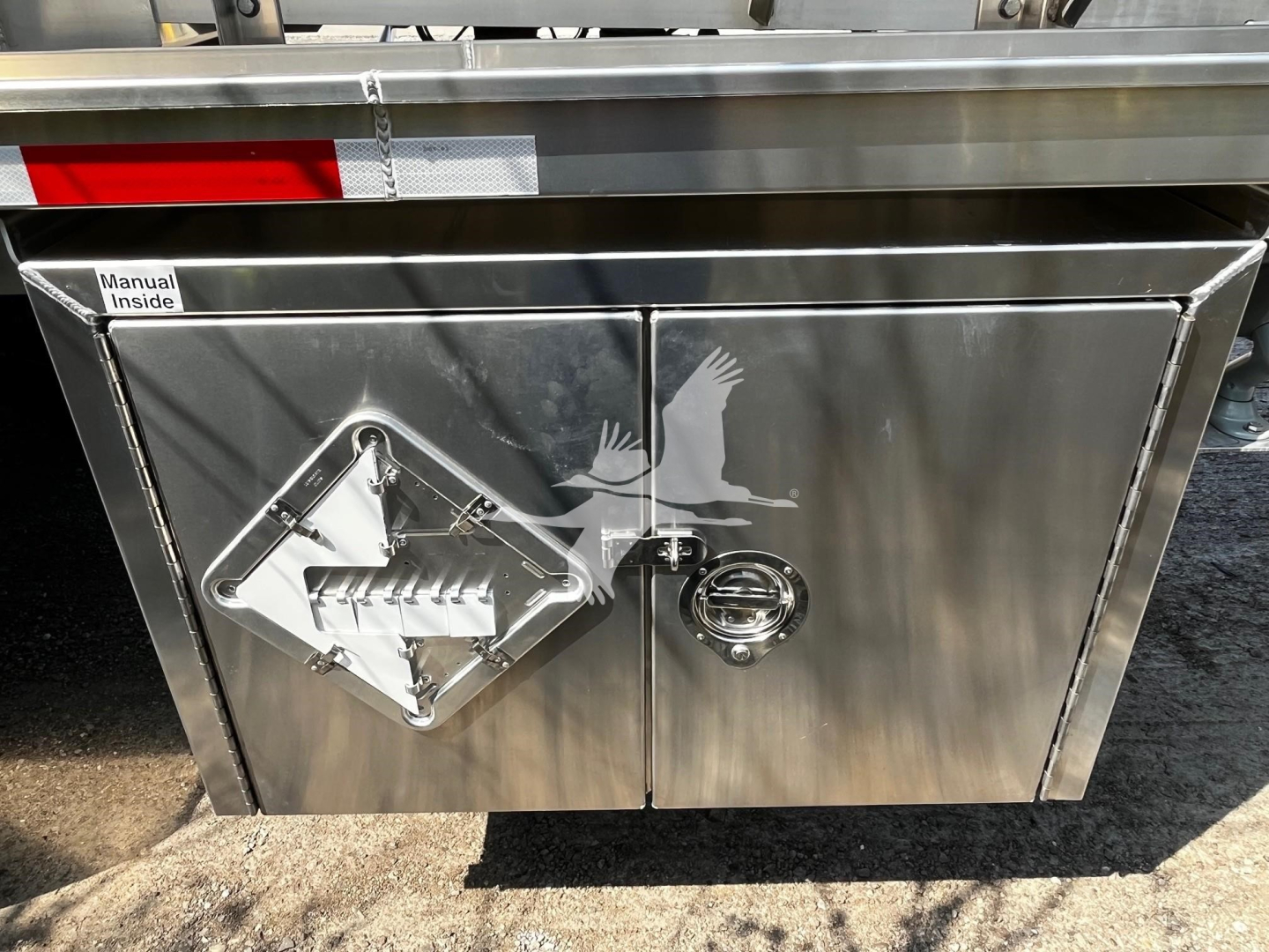 Trailer storage compartment door with diamond plate and gas can access hatch showing manual inside label.