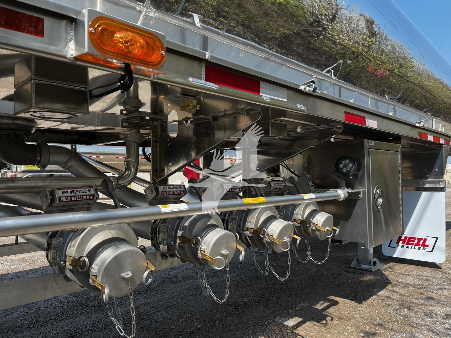 Trailer underside showing multiple couplings and hoses with protective chains and metal supports; Heil logo on rear wheel area and reflective lights.