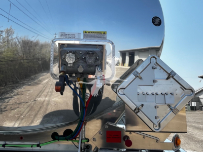 Trailer hitch area with control box and diamond-shaped access panel showing wiring harnesses, hoses, and metal frame.