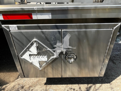 Trailer storage compartment door with diamond plate and gas can access hatch showing manual inside label.