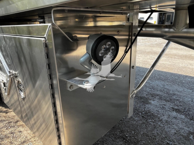 Quarter-view of a Heil trailer undercarriage with a round LED flood light mounted on a stainless panel, hydraulic lines and a silver toolbox visible.