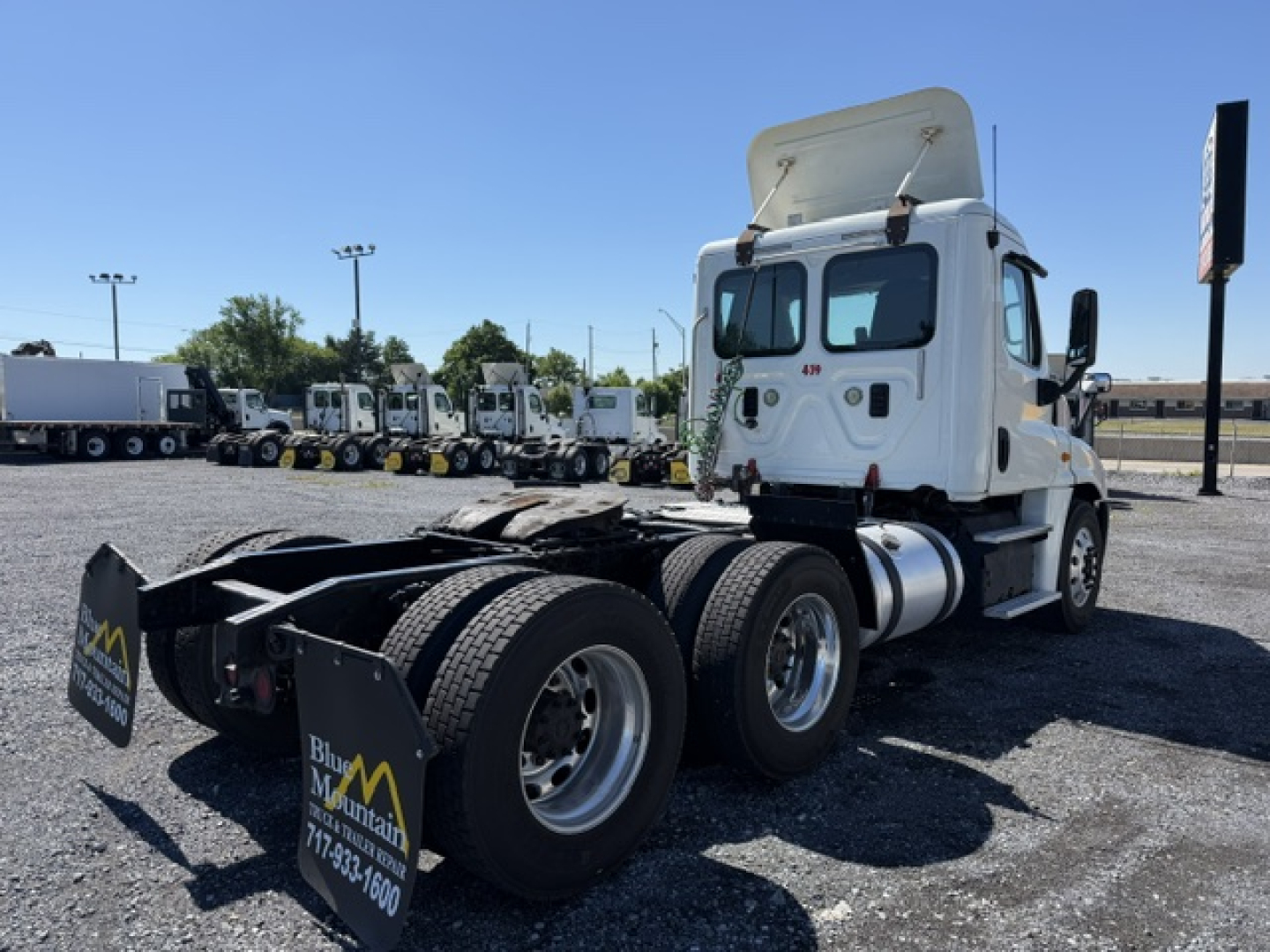 Freightliner Cascadia CA125DC tractor tractor rear view showing chassis and dual rear axles, white cab, raised sleeper door, fuel tanks, and parking lot with other trucks.