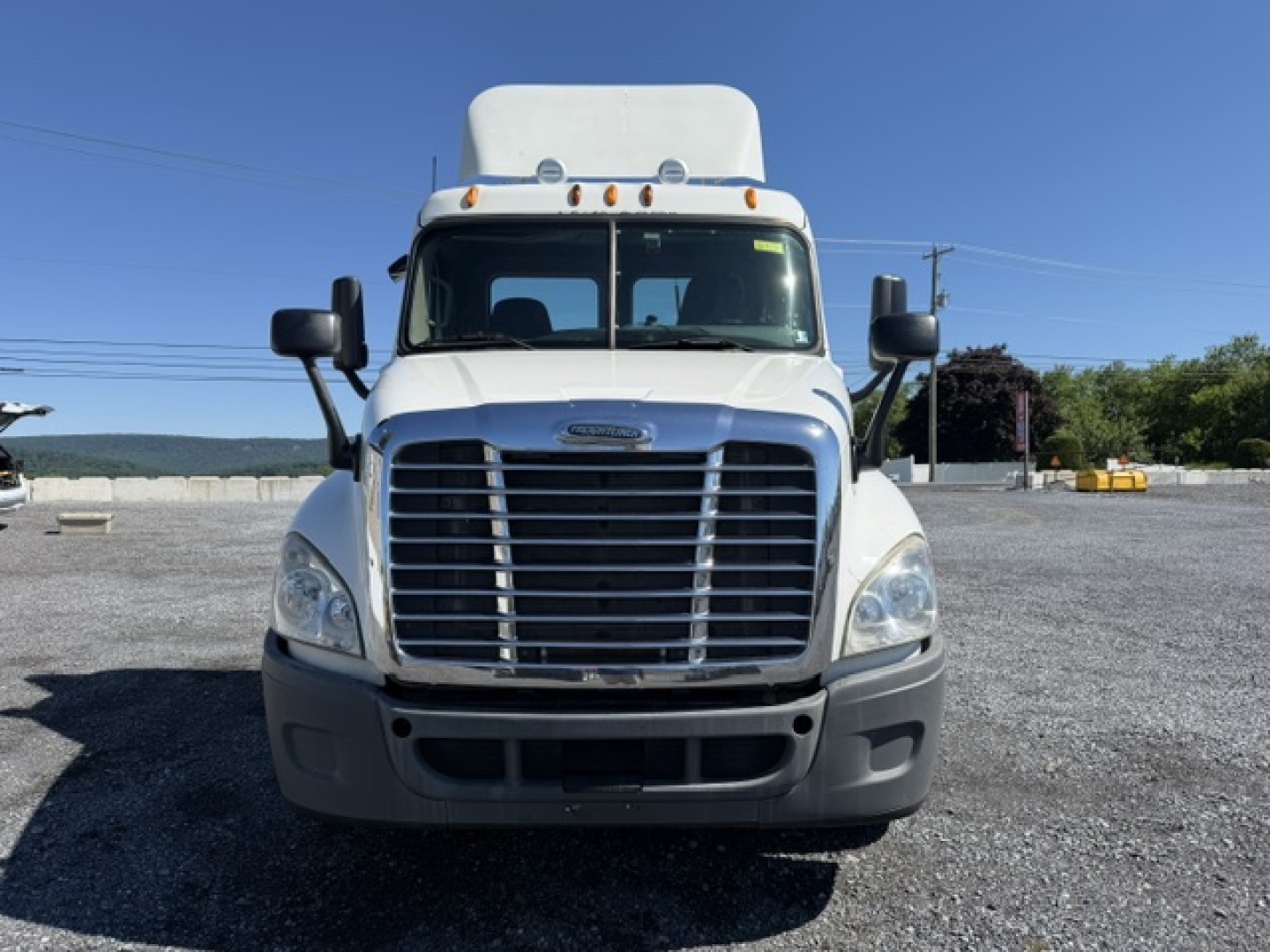 Front view of a white Freightliner Cascadia CA125DC truck in a lot with clear blue sky. Grille, headlights, bumper, and mirrors visible.