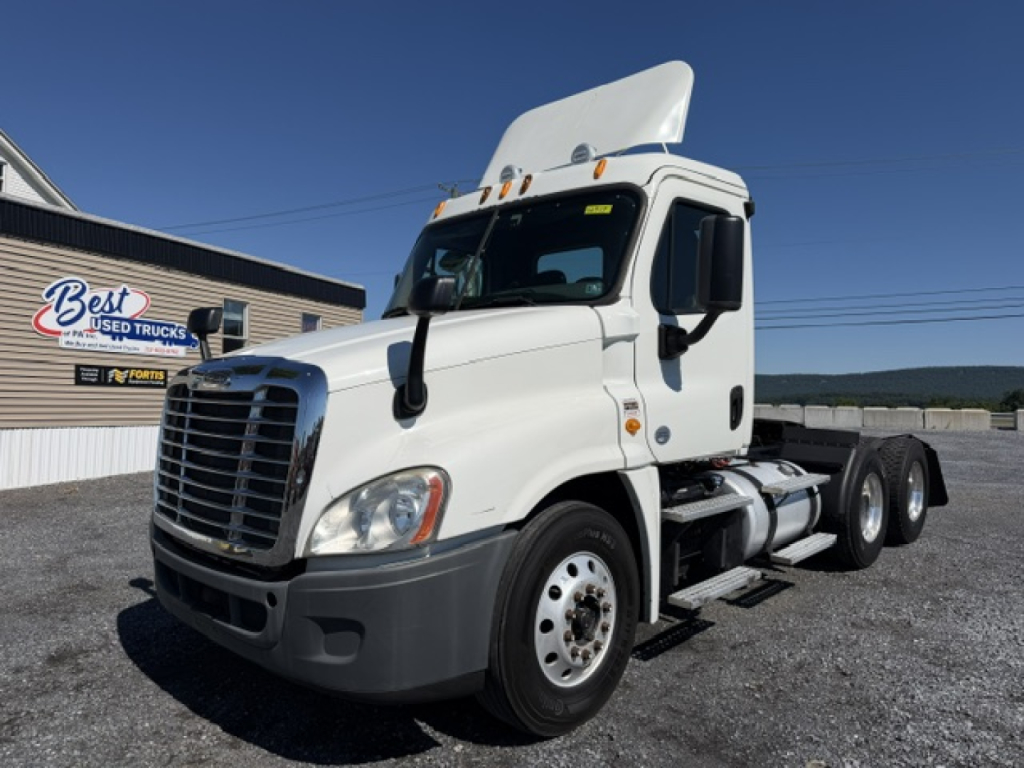 Truck cab in white with raised sleeper hood, Freightliner front grille, large mirrors, and fuel tanks visible. Text visible includes dealership sign in background reading 'Best Used Trucks' and a blue banner.