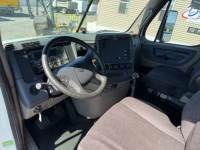 Cab interior of a Freightliner Cascadia dash and steering wheel with manual gear selector and gray/black upholstery.