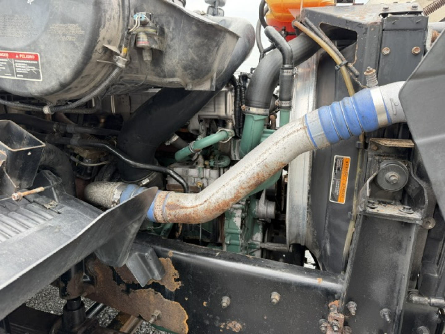 Truck engine bay showing radiator, hoses, and engine components with a prominent curved metal exhaust pipe and blue hose; some labels/stickers visible on components.