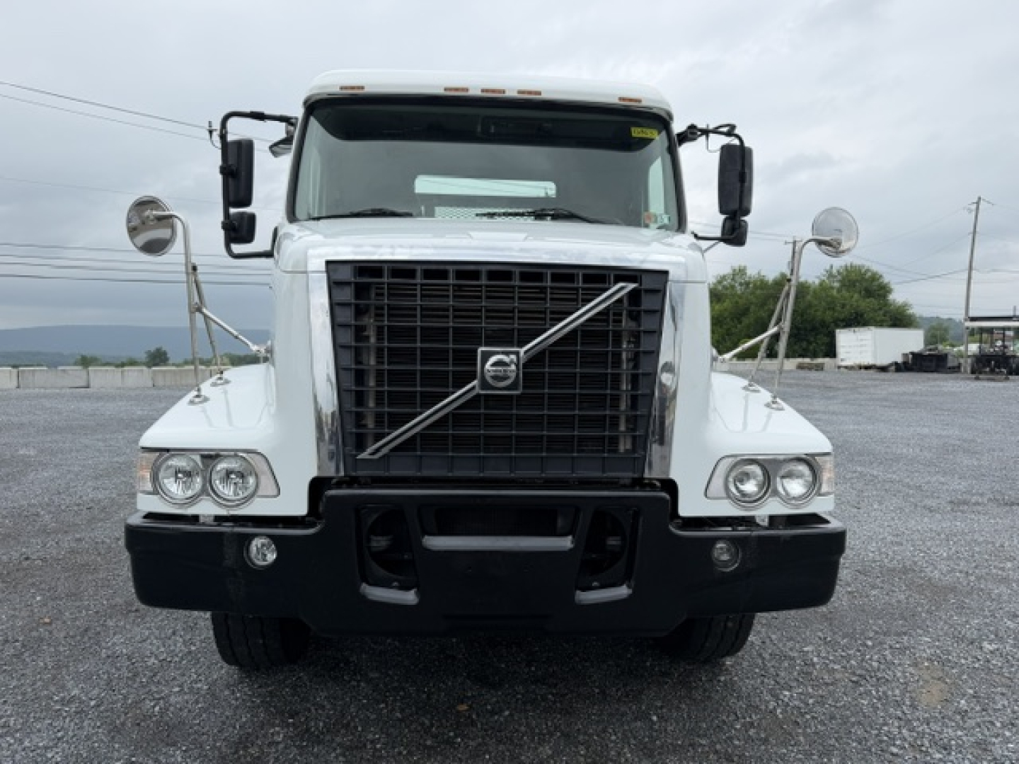 Volvo VHD front view showing large grille with Volvo emblem, dual rectangular headlights, black bumper, and side mirrors; flatbed truck aesthetic.